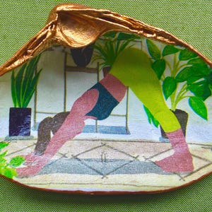 May include: A hand-painted seashell with a gold-painted rim and a yoga-themed interior. The artwork depicts a person in a downward-facing dog pose, surrounded by potted plants. The background is a light-colored room with a geometric floor pattern.