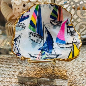 May include: A decorative, shell-shaped dish with a gold-painted edge. The dish features a nautical design with colorful sailboats on a white background. The sailboats have blue, pink, yellow, and green sails. The dish is displayed on a gold-colored metal stand.