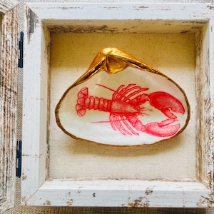 May include: A decorative seashell with a red lobster illustration, set within a distressed white wooden frame. The shell's rim is gilded with gold, and the background is a neutral linen fabric.