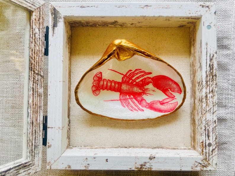 May include: A decorative seashell with a red lobster illustration, set within a distressed white wooden frame. The shell's rim is gilded with gold, and the background is a neutral linen fabric.