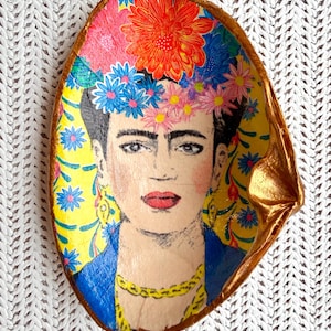 May include: A decorative shell dish featuring a hand-painted portrait of Frida Kahlo. The subject wears a vibrant floral crown with red, pink, and blue flowers against a yellow background with blue star-like patterns. The shell edge is painted gold.