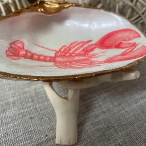 May include: A decorative seashell dish with a gold-painted rim and a red lobster illustration. The dish is supported by a small, off-white stand. The interior of the shell is white.