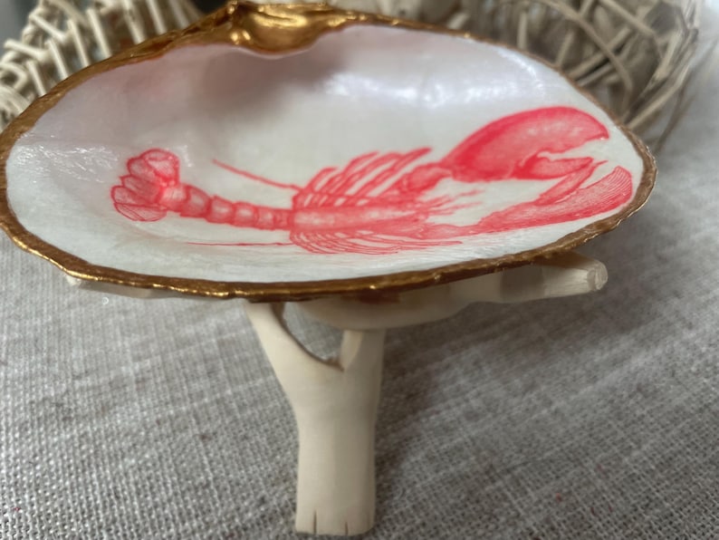 May include: A decorative seashell dish with a gold-painted rim and a red lobster illustration. The dish is supported by a small, off-white stand. The interior of the shell is white.