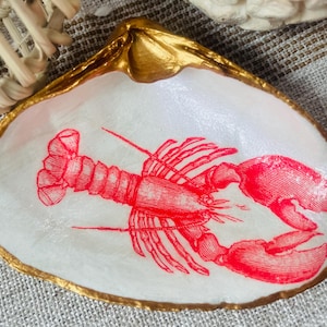 May include: A decorative seashell dish with a gold-painted rim. The interior features a red lobster illustration on a white background. The dish is likely used for holding small items.