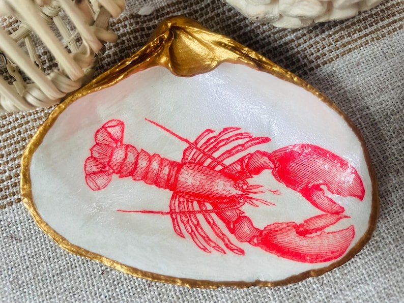 May include: A decorative seashell dish with a gold-painted rim. The interior features a red lobster illustration on a white background. The dish is likely used for holding small items.