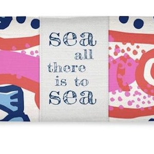 Puede incluir: Un cojín largo y rectangular con fondo crema. El cojín presenta un patrón repetitivo de peces y formas abstractas azules y rojas. El centro del cojín tiene el texto "sea all there is to sea".