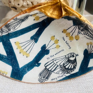 May include: Decorative seashell with a gold-painted edge. The shell features a white background with a blue tree branch design, yellow berries, and a black and white bird illustration. Text reads "You belong to the universe. -Mary Oliver."