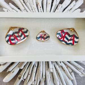 Puede incluir: Una obra de arte enmarcada en blanco con tres conchas pintadas. Las conchas tienen letras rojas y un patrón de chevron en blanco y negro. Las conchas están sobre un fondo blanco y rodeadas por un diseño de madera flotante.