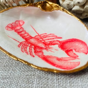 May include: A decorative seashell dish with a gold-painted rim. The white interior features a red lobster illustration. This coastal-themed dish is perfect for holding small items.