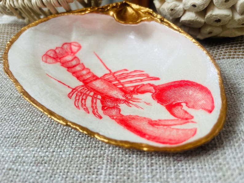 May include: A decorative seashell dish with a gold-painted rim. The white interior features a red lobster illustration. This coastal-themed dish is perfect for holding small items.