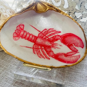 May include: A decorative seashell dish with a gold-painted rim and a red lobster illustration on a white background. The dish is resting on a neutral-colored surface.