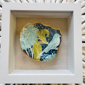 May include: A decorative, framed art piece featuring a blue bird and floral design. The artwork is set against a dark background with gold trim, encased in a white frame. The piece is displayed on a neutral linen-textured background.