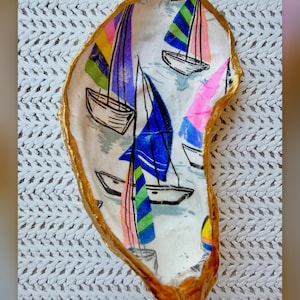May include: A decorative oyster shell dish featuring a colourful sailboat pattern with bright pink, blue, green, and yellow sails on a white background. The edges of the shell are painted with a metallic gold finish. The text @saltysisterdesign is visible.
