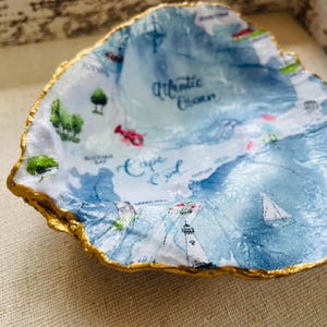 May include: A decorative, irregularly shaped dish with a map of Cape Cod printed on the interior. The map is in shades of blue and white, with a gold-painted rim. The dish features images of a lighthouse, sailboat, and lobster.
