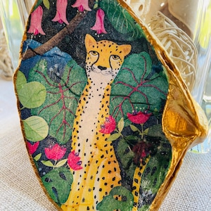 Op de afbeelding: Handbeschilderde decoratieve zeeschelp met een cheetah te midden van weelderig groen gebladerte en roze bloemen. De cheetah heeft gele vacht met zwarte vlekken. De rand van de schelp is verguld met goudverf, wat een uniek kunstwerk creëert.