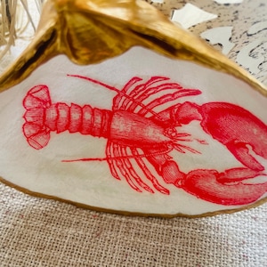 May include: A decorative seashell with a red lobster illustration. The lobster is detailed with fine lines and is centered on the white interior of the shell. The shell has a gold-colored rim and sits on a textured surface.