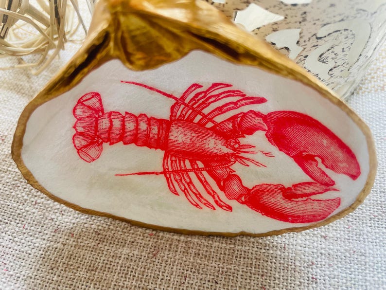 May include: A decorative seashell with a red lobster illustration. The lobster is detailed with fine lines and is centered on the white interior of the shell. The shell has a gold-colored rim and sits on a textured surface.