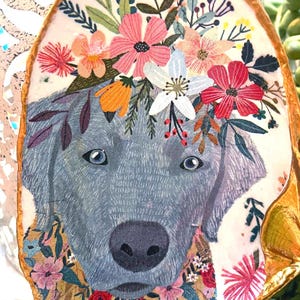 May include: Oval-shaped artwork featuring a gray dog with blue eyes, adorned with a floral crown of pink, white, and orange flowers. The dog's neck is surrounded by a floral pattern, set against a light background with a gold border.