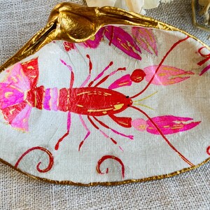 May include: A decorative seashell dish with a gold-painted rim and accents. The interior features a hand-painted red and pink lobster design on a cream-colored background. The dish is ideal for holding small items.