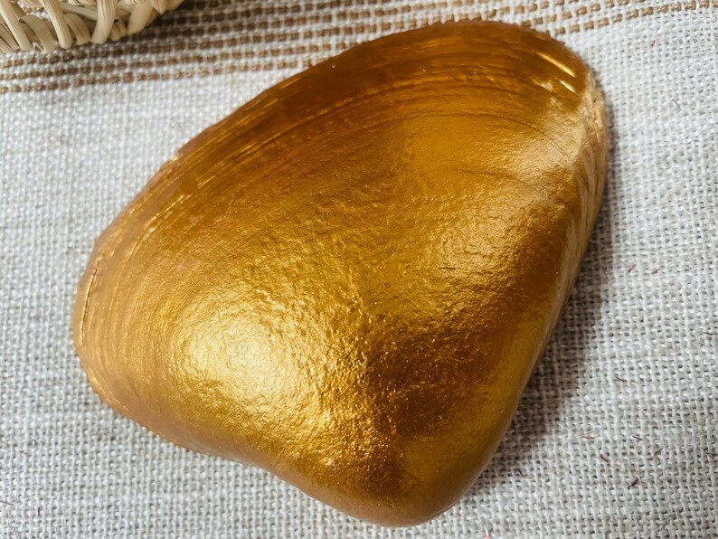 May include: A golden-colored, shell-shaped decorative object. The shell has a textured surface and is painted with a metallic gold finish. The shell is resting on a textured, light-colored fabric.