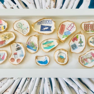 May include: A collection of decorative oyster shell dishes with gold rims, each hand-painted with a different Nantucket-themed design. The designs include a bicycle, lighthouse, sailboat, and the word "Nantucket".