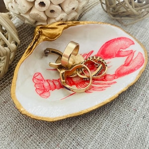 May include: A decorative seashell jewelry dish with a gold-painted rim and a red lobster illustration. The dish holds several gold rings and earrings. The dish is on a textured surface.