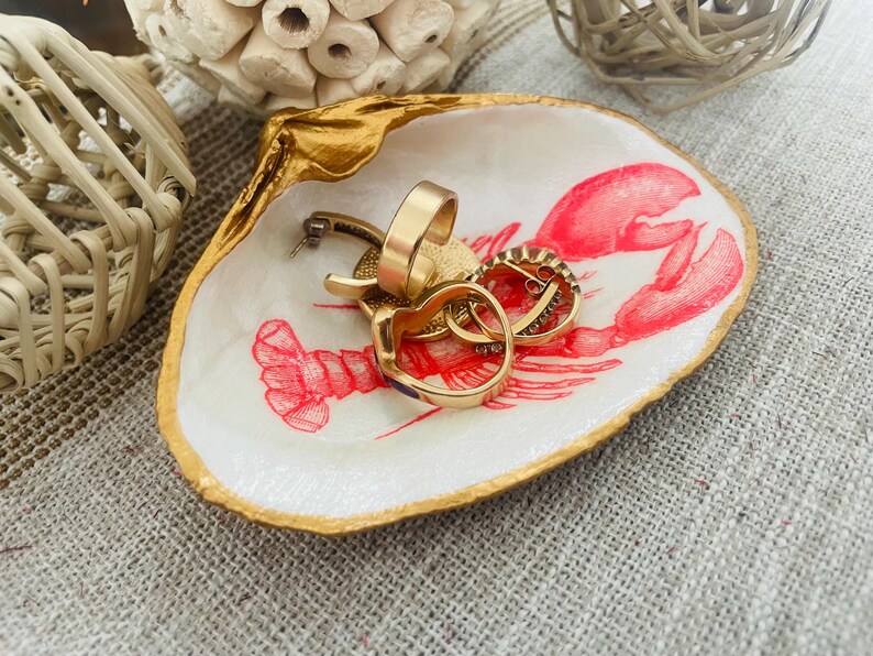 May include: A decorative seashell jewelry dish with a gold-painted rim and a red lobster illustration. The dish holds several gold rings and earrings. The dish is on a textured surface.