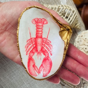 May include: A white seashell trinket dish with a red lobster illustration. The shell has a gold-painted rim and accents. The dish is held in a hand.