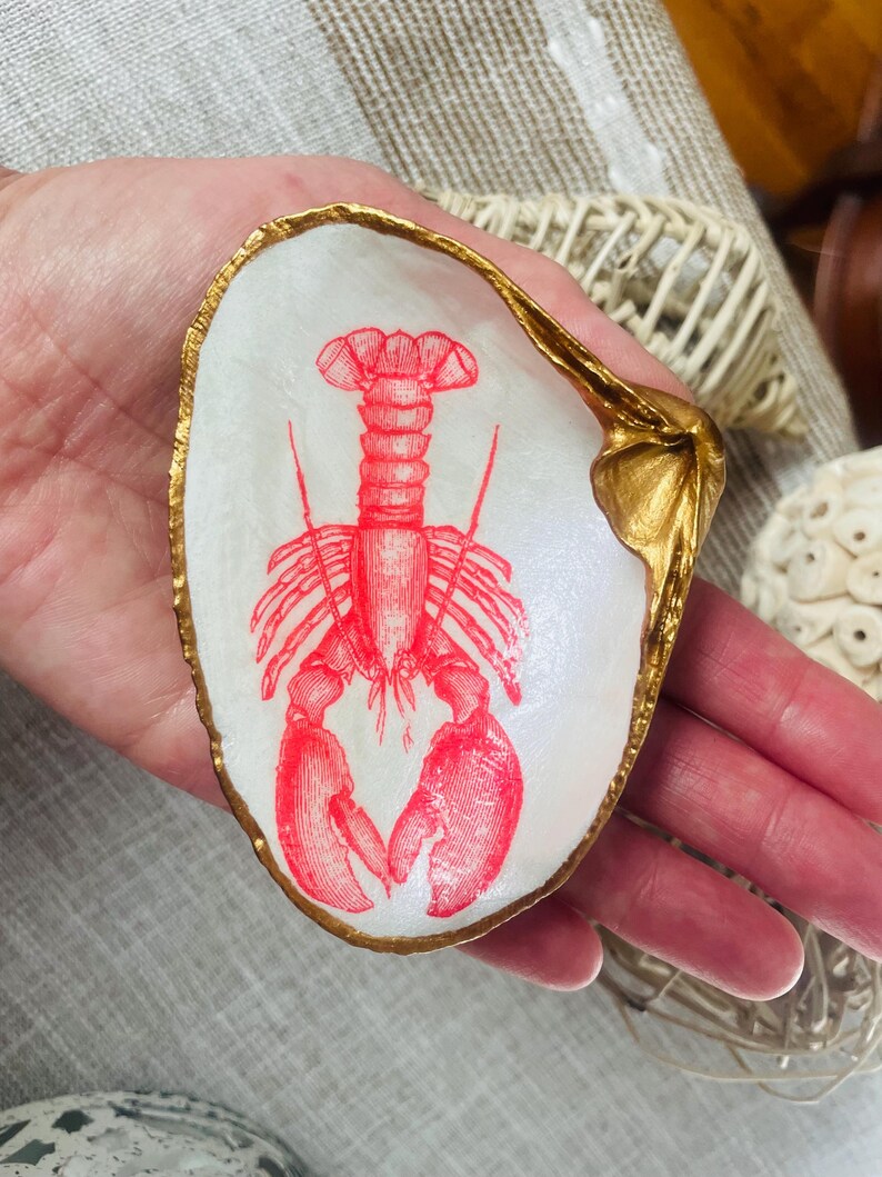 May include: A white seashell trinket dish with a red lobster illustration. The shell has a gold-painted rim and accents. The dish is held in a hand.