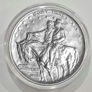 Puede incluir: Una moneda de plata que representa a dos hombres a caballo, con la inscripción "IN GOD WE TRUST" en la parte superior. La moneda está inscrita con "STONE MOUNTAIN" y el año "1995". La moneda está dentro de una funda protectora transparente.