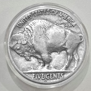 May include: A silver Buffalo nickel coin encased in a clear plastic holder. The coin features a detailed profile of a bison on the right side and the words "UNITED STATES OF AMERICA" and "FIVE CENTS" around the edges.