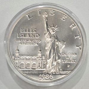 May include: A silver coin featuring the Statue of Liberty, Ellis Island, and the text "Gateway to America." The coin is dated 1986 and includes the inscription "In God We Trust."