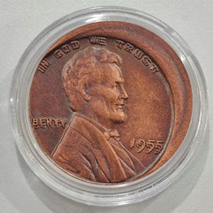 Może przedstawiać: Miedziana moneta Lincoln z 1955 roku w przezroczystym plastikowym etui. Moneta przedstawia profil Abrahama Lincolna oraz napisy "IN GOD WE TRUST" i "LIBERTY". Widoczny jest również rok "1955".