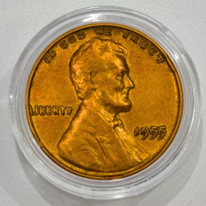 Op de afbeelding: Een 1955 Lincoln penny in een doorzichtige beschermhoes. De koperen munt toont een profiel van Abraham Lincoln, met de woorden "LIBERTY" en "IN GOD WE TRUST" zichtbaar. Het jaartal "1955" is ook weergegeven.