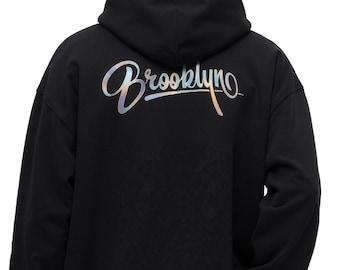 May include: Black hoodie with a drawstring hood and long sleeves. The word "Brooklyn" is printed on the back in a holographic, cursive font. The hoodie is made of a soft material and has a relaxed fit.