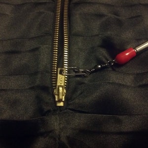 May include: A gold zipper is pulled open on a black fabric garment. A safety pin is attached to the zipper pull and connected to a red and silver fishing lure.