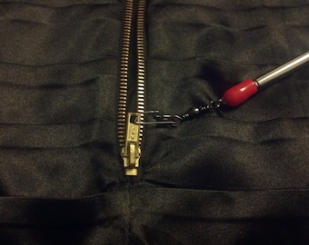 May include: A gold zipper is pulled open on a black fabric garment. A safety pin is attached to the zipper pull and connected to a red and silver fishing lure.