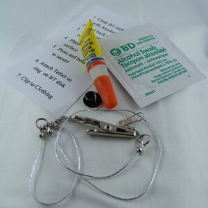 May include: A kit containing a yellow and orange adhesive tube, an alcohol swab, a small black disk, and a silver cord with a clip. Instructions are printed on a white paper. The kit is designed for a specific purpose.