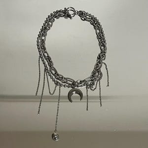 Op de afbeelding: Een zilverkleurige ketting met meerdere lagen en een centrale halve maan hanger. Het ontwerp combineert dikke ovale schakels en delicate kralenkettingen, met verschillende dunne bungelende kettinkjes en een kleine bolvormige bedel.