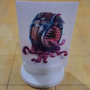 May include: A white game piece with a monster illustration. The monster has a barrel-like body, blue skin, and a large mouth with a pink tongue. The piece is on a grid-patterned surface.
