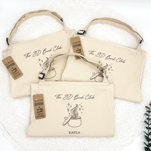 May include: Off-white canvas aprons with adjustable straps. Each apron features the text "The SD Book Club" and a graphic of a pot with steam. One apron is personalized with the name "KAYLA". Each apron has a tag that says "THE ARTISAN REPRIME APRONS".