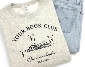 May include: A heather grey sweatshirt with the text "Your Book Club" and a stylized image of an open book with stars above it. The text "One more chapter" and "EST. 2025" are below the book image.