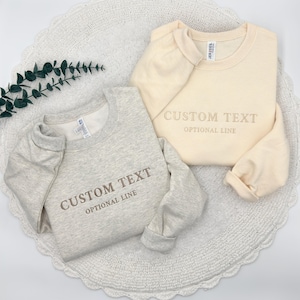 May include: A light gray heather sweatshirt and a cream-colored sweatshirt, both featuring embroidered text that reads CUSTOM TEXT and OPTIONAL LINE. Each crewneck garment has long sleeves and a visible brand tag inside the collar area.