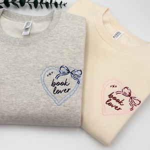 May include: A light gray heather sweatshirt and a cream sweatshirt, each featuring an embroidered heart-shaped design with a bow and the text "book lover". The gray sweatshirt has blue embroidery, while the cream sweatshirt has pink embroidery.