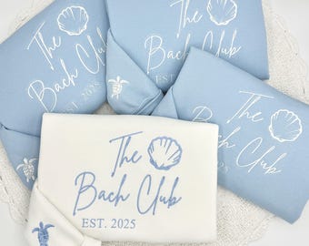 May include: A collection of light blue and white sweatshirts and a matching face mask. The sweatshirts feature the text "The Bach Club" with a seashell graphic and "EST. 2025". The face mask has a turtle graphic.