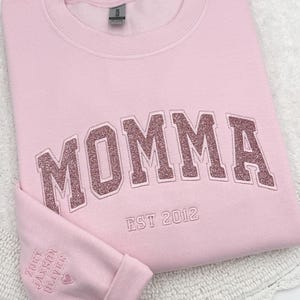 May include: Pink sweatshirt with the word "MOMMA" in pink glitter lettering and the text "EST 2012" below. The sleeve has the names "ZOBY, JACKSON, OLIVER" and a heart.