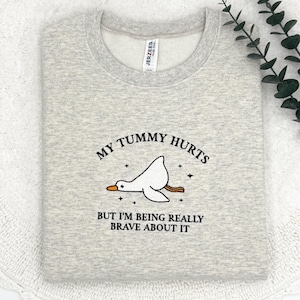 May include: A gray sweatshirt with the text "My tummy hurts but I'm being really brave about it" and a cartoon illustration of a goose with its head tilted back.