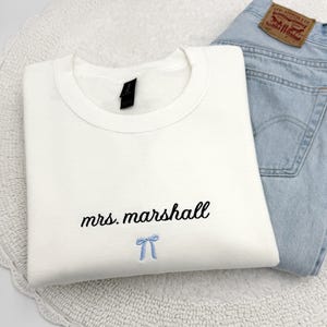 May include: A white crew neck sweatshirt with the embroidered text "mrs. marshall" in black script and a light blue bow. Light blue denim jeans are visible in the background.