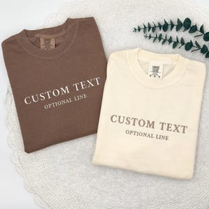 May include: A brown t-shirt and a cream t-shirt, both featuring the printed text CUSTOM TEXT and OPTIONAL LINE on the front. Each shirt has a crew neckline and a Comfort Colors brand label visible on the inside of the collar area.