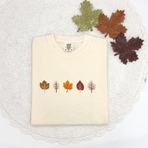 May include: Cream-colored t-shirt with embroidered fall leaves in shades of brown, orange, and pink. The shirt is folded neatly on a white lace doily, with glittery leaf decorations in the background. The shirt has a Comfort Colors tag.